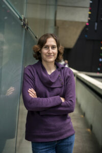 Katherine Mack, wearing a purple cowl-necked sweater standing in a hallway